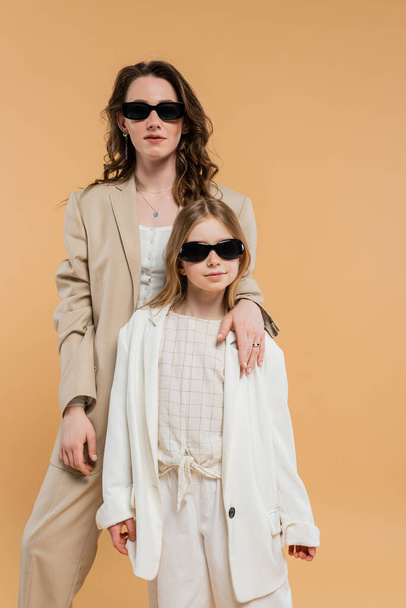 modern gezin, stijlvolle moeder en dochter in zonnebril, zakenvrouw en meisje in pakken die samen op beige achtergrond staan, modieuze outfits, formele kledij, huismoeder  - Foto, afbeelding