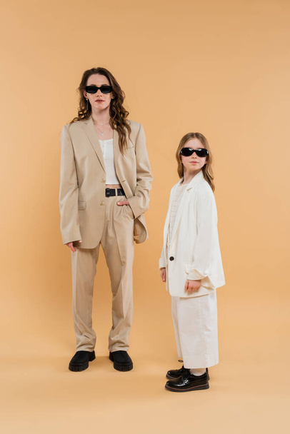 nowoczesna rodzina, matka i córka w okularach przeciwsłonecznych, stylowa businesswoman i dziewczyna w garniturach pozujących razem na beżowym tle, modne stroje, strój formalny, mama korporacyjna  - Zdjęcie, obraz