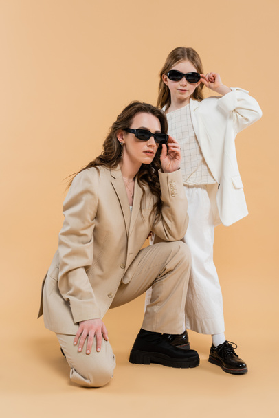 modern family, stylish mother and daughter in suits and sunglasses, businesswoman sitting near girl on beige background, fashionable outfits, formal attire, working mother, trendsetter  - 写真・画像