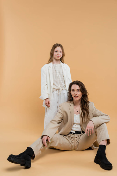 corporate mom concept, stylish woman in suit sitting near preteen daughter and looking at camera on beige background, togetherness businesswoman, motherly love  - Fotografie, Obrázek