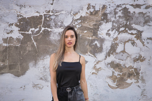 Young, pretty, blonde woman in black tank top and jeans on a wall with sagging paint, looking at camera self-confident. Self-confident concept, confidence, security. - Фото, зображення