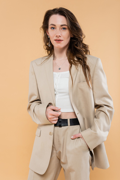 fashion trend concept, young woman with wavy hair standing in fashionable suit and looking at camera on beige background, hand in pocket, classic style, stylish posing  - Foto, imagen