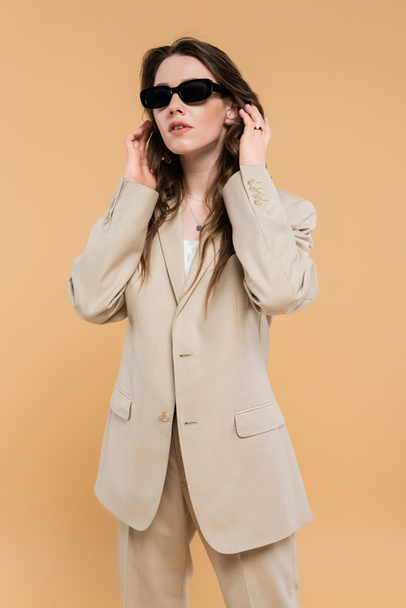fashion trend concept, young woman adjusting wavy hair and standing in fashionable suit with sunglasses on beige background, classic style, chic stylish posing, professional attire  - Фото, зображення