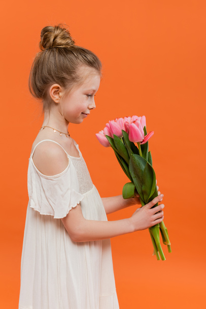 vista lateral de niña preadolescente en vestido de sol blanco con tulipanes rosados sobre fondo naranja, concepto de moda y estilo, ramo de flores, niño de moda, colores vibrantes, flores y moda  - Foto, imagen
