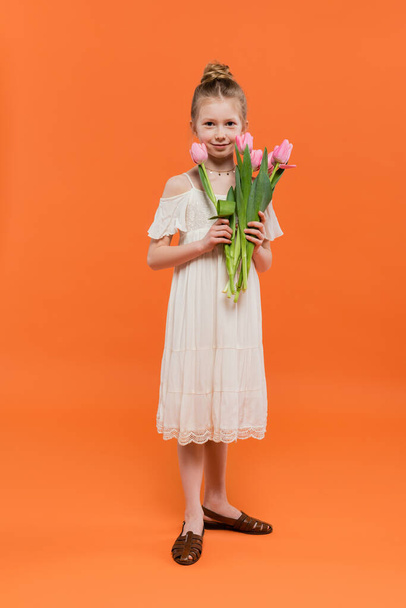 élénk színek, vidám tizenéves lány fehér napfényben ruha kezében rózsaszín tulipán narancssárga háttér, divat és stílus koncepció, csokor virágok, divatos gyerek, teljes hossz  - Fotó, kép