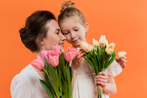 happy mother and daughter with tulips, young woman and girl holding flowers and posing on orange background, summer fashion, sun dresses, female bonding, family love  - Foto, imagen