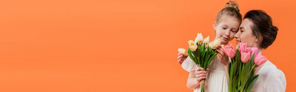 joyful mother and daughter with tulips, young woman and girl holding flowers and posing on orange background, summer fashion, sun dresses, female bonding, family love, banner  - Photo, Image
