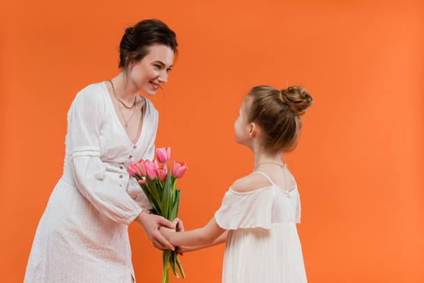 Mother`s day, preteen girl giving bouquet of flowers to smiling mother on orange background, bonding, white dresses, pink tulips, happy holiday, vibrant colors, joyful occasion  - Photo, Image