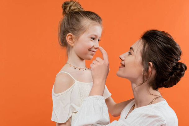 family bonding, cheerful mother touching nose of preteen daughter on orange background, white sun dresses, modern parenting, summer fashion, togetherness, love, fashionable family  - Foto, Bild