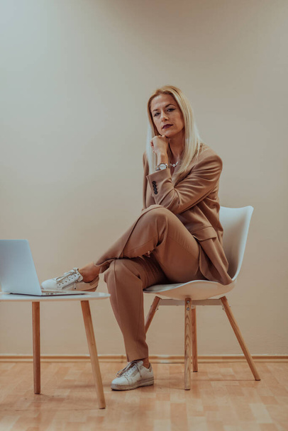 A professional businesswoman sits on a chair, surrounded by a serene beige background, diligently working on her laptop, showcasing dedication and focus in her pursuit of success. - Фото, зображення