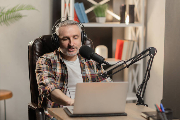 Starší muž ve sluchátkách dělá podcast. Blogger nahrává video s profesionálním mikrofonem u stolu. Kvalitní fotografie - Fotografie, Obrázek