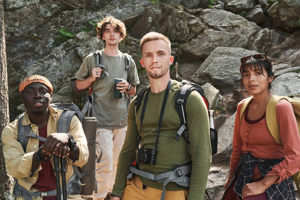 岩の上に立ってカメラを見ている袋を持つ自信に満ちた若い多民族ハイカーのグループ - 写真・画像