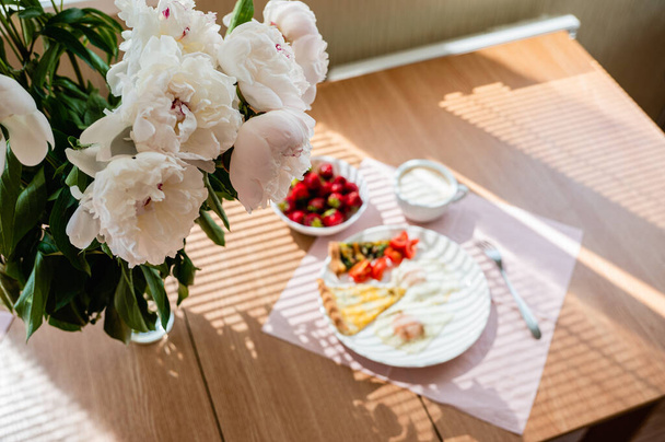 Fried eggs with a slice of pizza and a cup of cappuccino on the table. A beautiful bouquet of peonies stands in a vase on the table. Breakfast concept. - Photo, Image