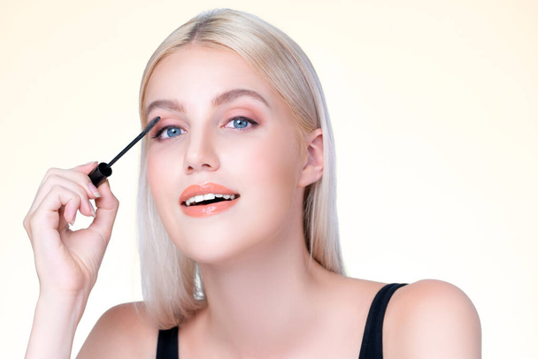 Closeup personable woman with blond hair putting black mascara with brush in hand on long thick eyelash. Perfect fashionable cosmetic clean facial skin with beautiful eye young woman. - Photo, Image