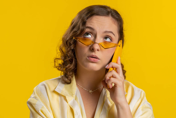 Tired sleepy young woman in glasses talking on mobile phone with friend making online conversation. Disinterested attractive girl having annoyed boring talk on smartphone isolated on yellow background - Photo, Image