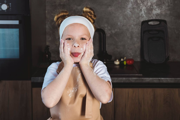 Cooking or Culinary school for children, kids. Little girl with hands and a face smeared in flour, having fun, cooks on the kitchen and fooling around. - Photo, Image