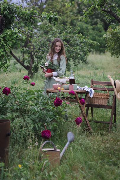 girl serves a table for tea drinking in the garden and in the foreground a bush of peonie - Fotografie, Obrázek