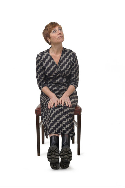 Vorderseite eine Frau in Kleid und Stiefeln, die auf einem Stuhl sitzt und auf weißem Hintergrund nach oben schaut - Foto, Bild