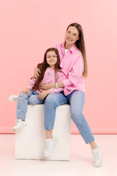 A young mother has fun playing with a cute little daughter, holding in her arms, gently hugging on a pink background - Photo, Image