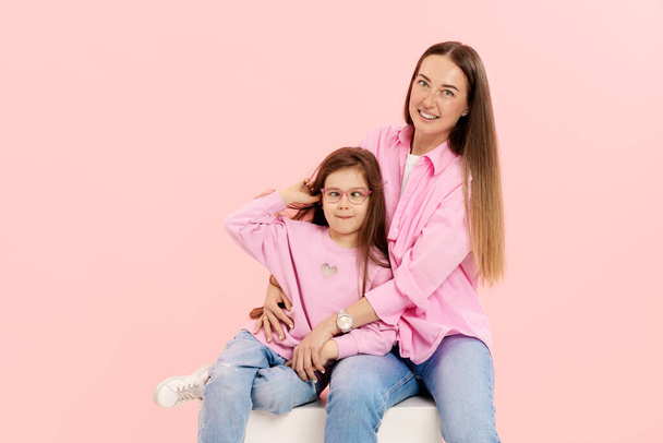 A young mother has fun playing with a cute little daughter, holding in her arms, gently hugging on a pink background - Фото, изображение