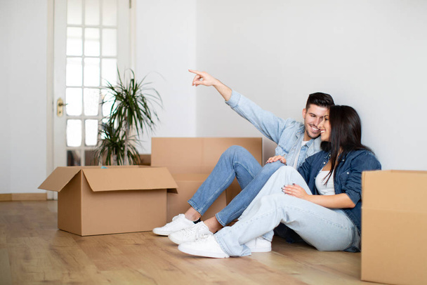 Young Happy Couple Planning Interior Design In Their New Home After Moving, Smiling European Spouses Sitting On Floor Among Cardboard Boxes And Pointing Away, Imagining Decorations And Furniture - Photo, Image