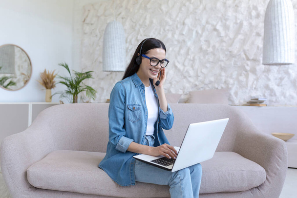 A young female student studies at home remotely on a laptop. He is sitting on the couch in a headset and jeans. Talks via video call, zoom meeting, listens to online lectures and lessons. - Photo, image