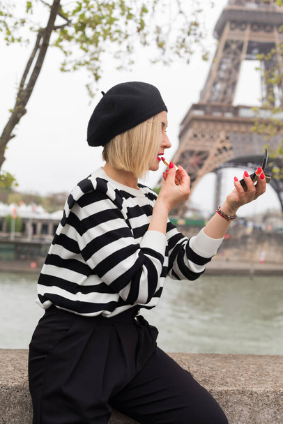 Charming female with short blonde hair painting lips with bright red lipstick while sitting on stone fence. Woman in striped black and white blazer primping against backdrop of Eiffel Tower. - Foto, immagini