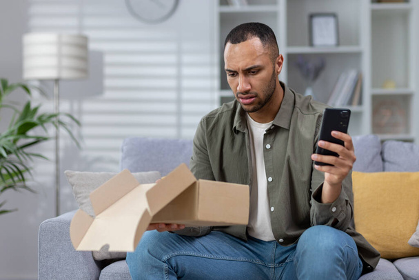 A worried young Hispanic man received a wrong and damaged package, an online store order, at home. He sits at home on the sofa with the phone and contacts the post office, the delivery service. - Φωτογραφία, εικόνα