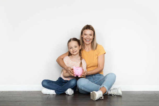 Rodinný rozpočet. Usmívající se krásná matka a malá dcera drží prasátko banky při relaxaci na podlaze u bílé zdi, šťastný maminka a žena dítě těší ekonomiku a peníze úspory, kopírovat prostor - Fotografie, Obrázek