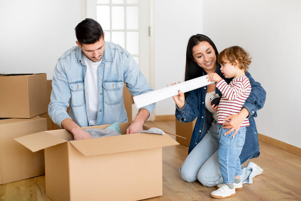 Концепция дня переселения. Веселая семья из трех распаковывающих картонную коробку с вещами в новом доме, счастливые молодые родители и милый малыш сын переезжает в свою квартиру или дом, крупным планом - Фото, изображение
