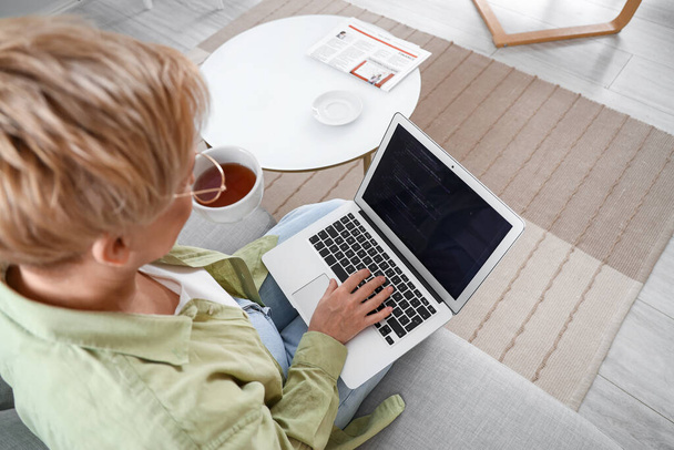 Kypsä naisohjelmoija työskentelee kannettavan tietokoneen ja kupin teetä sohvalla kotona, lähikuva - Valokuva, kuva