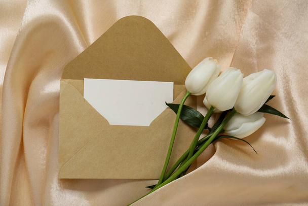 Φάκελος με λευκή κάρτα πρόσκλησης και όμορφα λουλούδια τουλίπας σε μπεζ τσαλακωμένο τραπεζομάντηλο - Φωτογραφία, εικόνα
