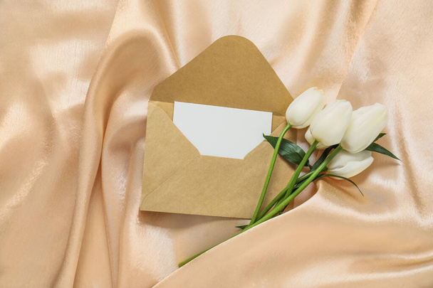 Φάκελος με λευκή κάρτα πρόσκλησης και όμορφα λουλούδια τουλίπας σε μπεζ τσαλακωμένο τραπεζομάντηλο - Φωτογραφία, εικόνα