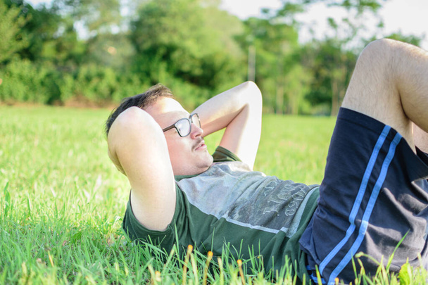Młody człowiek w okularach i zielonej koszulce robi przysiady i ćwiczy w parku z trawą i słonecznym dniem - Zdjęcie, obraz