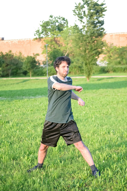 Молодой человек в зеленой футболке тренируется и растягивается в парке с травой и солнечным днем - Фото, изображение