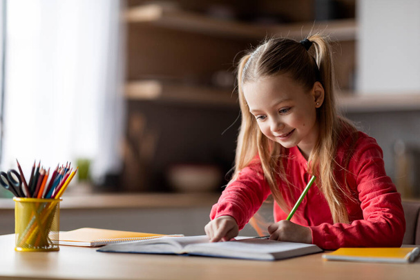 Concepto de Educación. Linda colegiala preadolescente haciendo tarea, Sonriendo niña pequeña escribiendo en el libro de trabajo mientras está sentada en el escritorio en el interior de la cocina, Comprobación de tarea en el libro, Copiar espacio - Foto, imagen