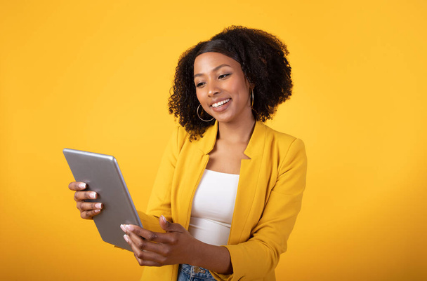 Cheerful black lady student typing on tablet, watching video and smiling isolated on yellow background, studio. Device and app for freelance, work and study, ad and offer, online sale - Valokuva, kuva