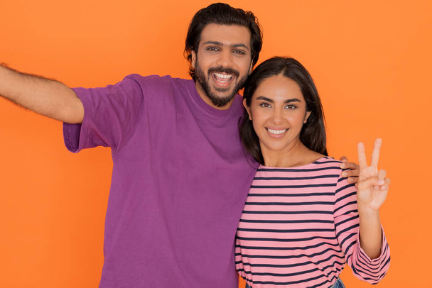 Happy loving beautiful eastern young couple wearing casual outfit taking selfie together, pretty indian woman showing peace gesture, isolated on orange studio background - Photo, Image