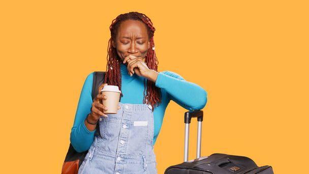 疲れ切った女性はコーヒーカップを飲み、スタジオであくびをし、飛行機で休暇で旅行する準備をしています。旅行者は眠くなり、カフェイン飲料を提供しています。手持ち撮影. - 写真・画像