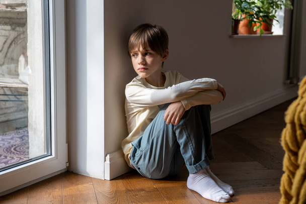 Ψυχολογικό τραύμα παιδιού. Μοναχικό παιδί δυστυχώς κοιτάζει στα γόνατα αγκαλιά παράθυρο κάθεται στο πάτωμα στο σπίτι μόνο. Αναστατωμένο προσβεβλημένο παιδί που σκέφτεται οικογενειακά προβλήματα, κακή σχέση, αποτυχίες με συμμαθητές στο σχολείο - Φωτογραφία, εικόνα