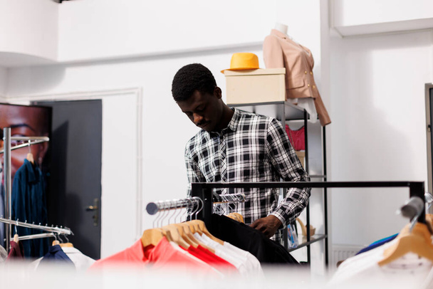 Homme afro-américain vérifiant des vêtements élégants dans une boutique moderne, faisant du shopping pour des marchandises à la mode. Élégant client analysant le matériel de produits et le prix dans le magasin de vêtements. Concept de mode - Photo, image