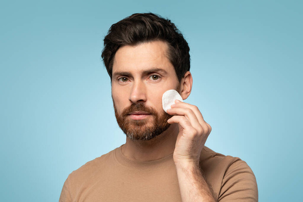 Soothing care. Mans relaxing skincare ritual with toner and cotton pad, looking at camera, posing over blue studio background. Beauty and skin care concept - Zdjęcie, obraz