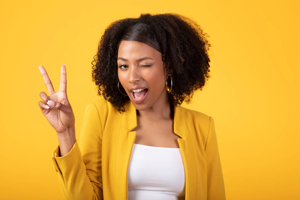 Vreugdevolle zwarte krullende vrouw knipoogt en toont vrede teken met de hand, poseren geïsoleerd op gele achtergrond, studio schot. Gesture, advertentie en aanbod, fashion blog, beauty care, positieve levensstijl - Foto, afbeelding