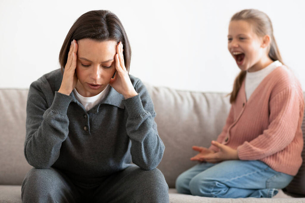 Unhappy caucasian middle aged mother suffers from screams and whims of daughter in living room interior. Relationship problems at home, parenting, scandal and stress with troubled teenager - Photo, Image