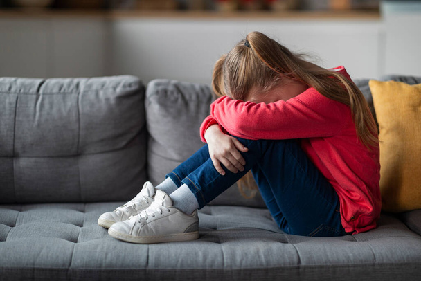 Kids Depression Concept. Upset Little Girl Crying At Home, Depressed Female Child Burying Head In Knees While Sitting On Couch In Living Room, Suffering Mental Breakdown, Copy Space - Photo, Image