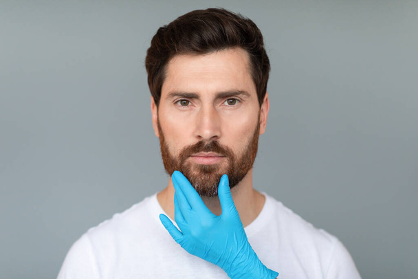 Szakértő. Kozmetikus kezek védő orvosi kesztyű érinti középkorú férfi arcát szürke stúdió háttér. Plasztikai sebészet és esztétikai kozmetikai koncepció - Fotó, kép