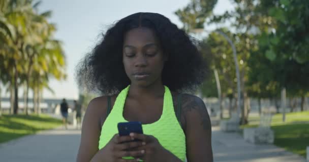 Lassú felvétel egy afro-amerikai fiatal nőről, aki SMS-ezik a mobilján, miközben a parkban van. - Felvétel, videó