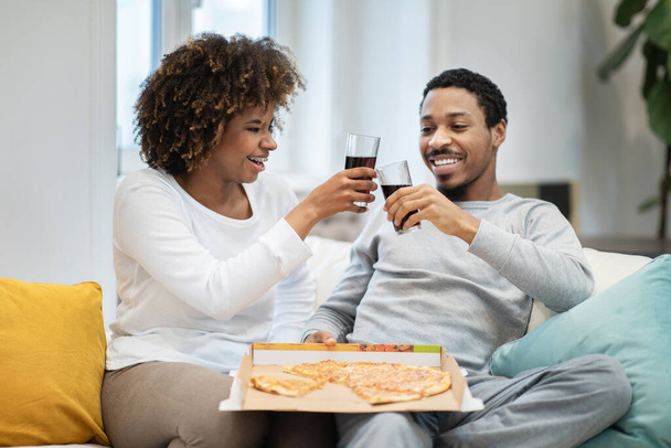 Cheerfy beaux jeunes amateurs noirs ont dîner à la maison. Heureux homme et femme afro-américain en tenue de maison assis sur le canapé, manger de la pizza, acclamer avec des lunettes, boire, célébrer bonne journée - Photo, image