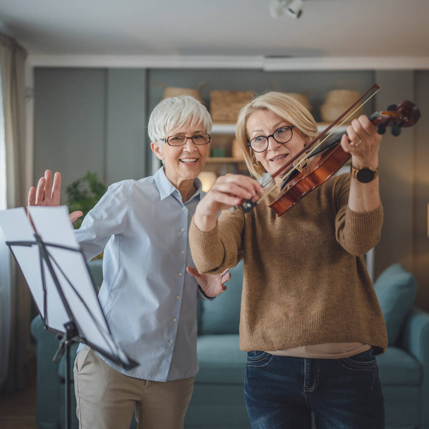 Two women mature female teacher give private violin lesson to her student or friend at home help her to learn to play in room hobby and leisure active senior concept copy space - Photo, Image