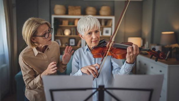Two women mature female teacher give private violin lesson to her student or friend at home help her to learn to play in room hobby and leisure active senior concept copy space - Photo, Image